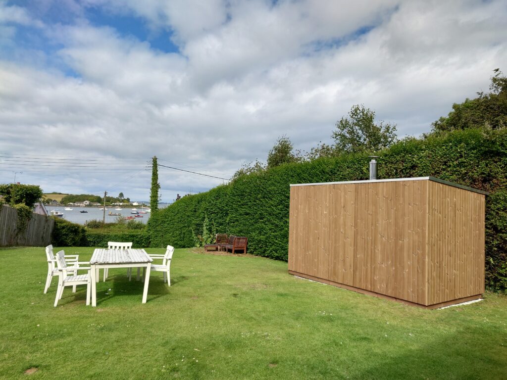 A timber outdoor sauna sited against a tall hedge for shelter at the edge of a lawn, with garden furniture nearby.