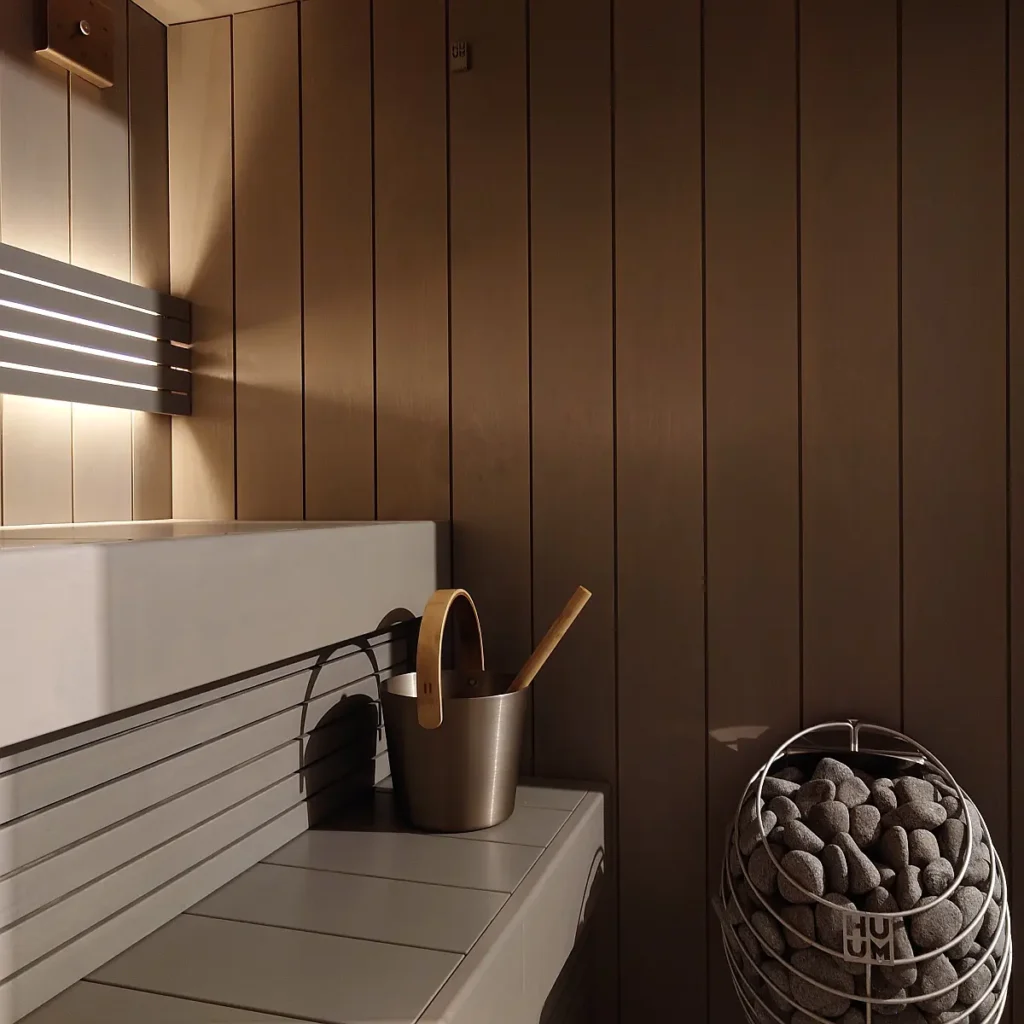 Interior of a modern indoor home sauna with dark wood panelling, a heater filled with sauna stones and a bench.