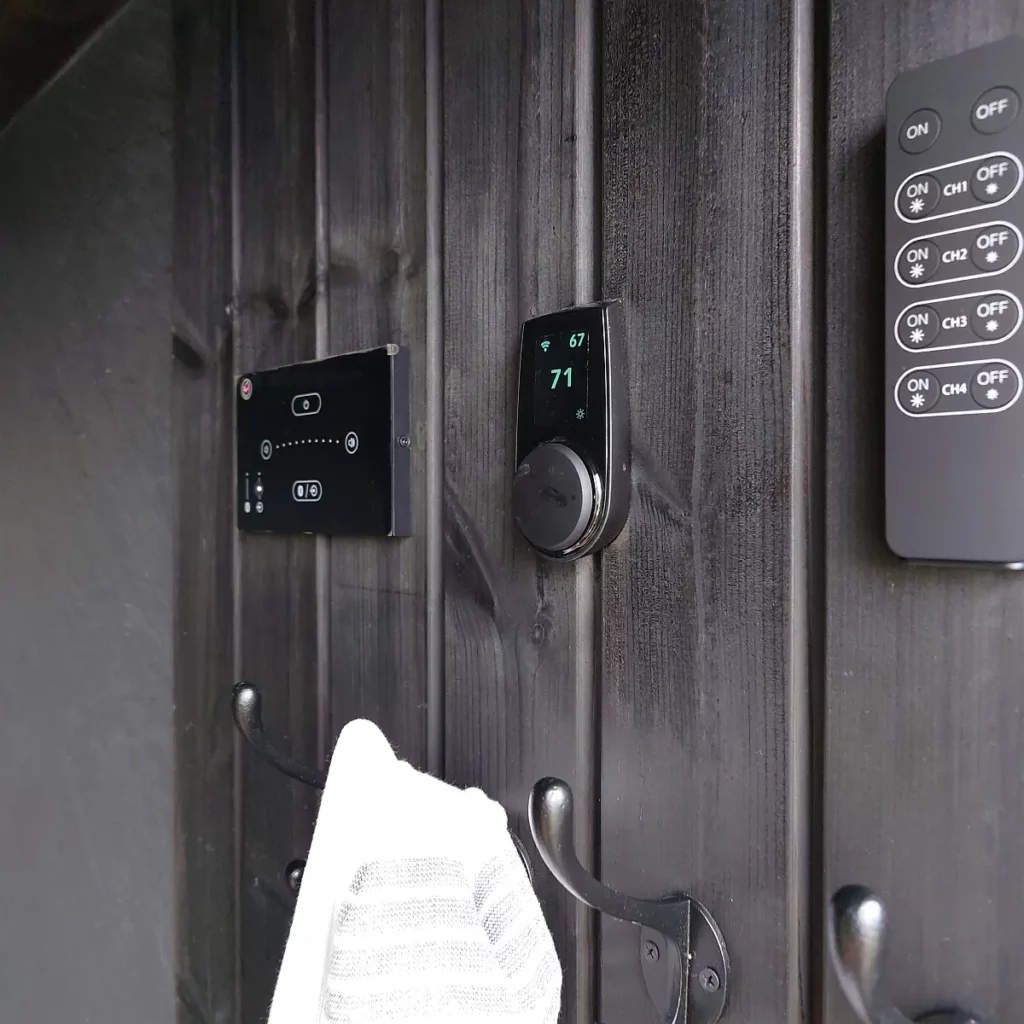 Digital sauna heater controls mounted on dark timber cladding beside a sauna, with towel hooks below.