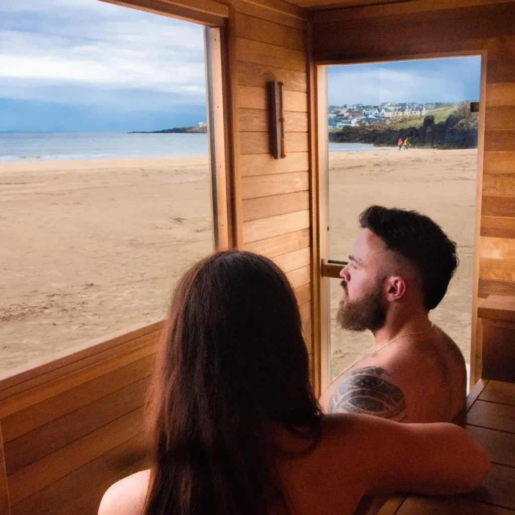 Two people relaxing inside a wooden sauna with a large window overlooking a sandy beach and coastline.