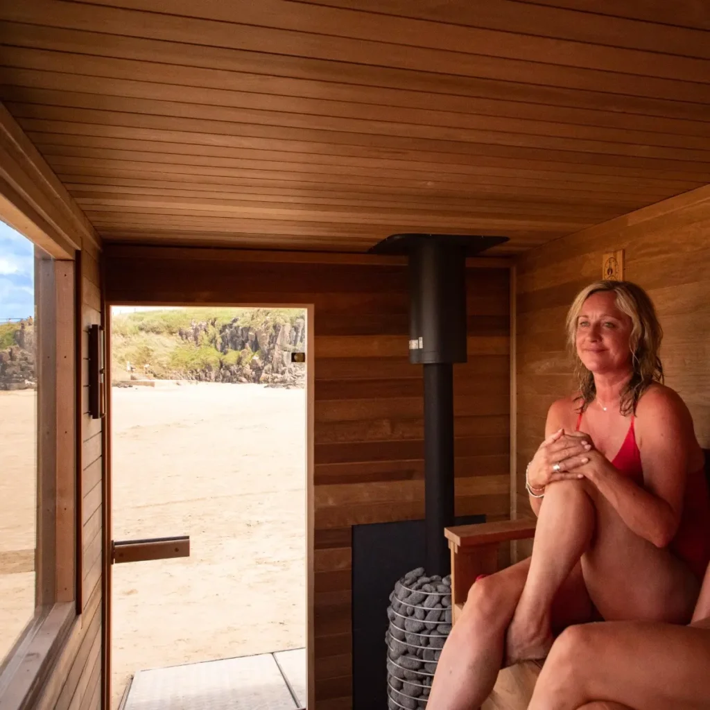 A woman in a swimsuit relaxing on the bench of a wooden sauna, with the door open to a sandy beach.
