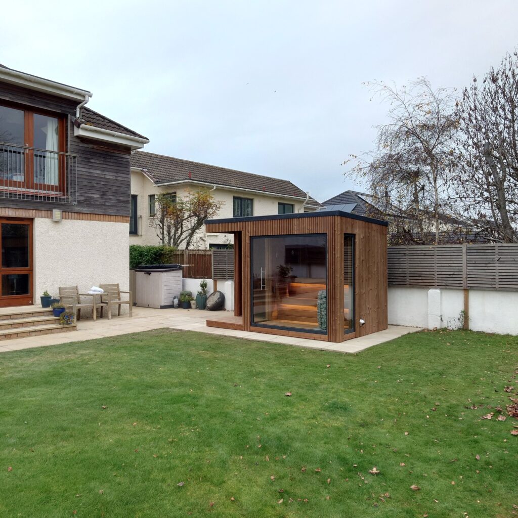 Modern freestanding wooden outdoor sauna with large glass front, positioned on a paved area in a landscaped garden beside a contemporary house.